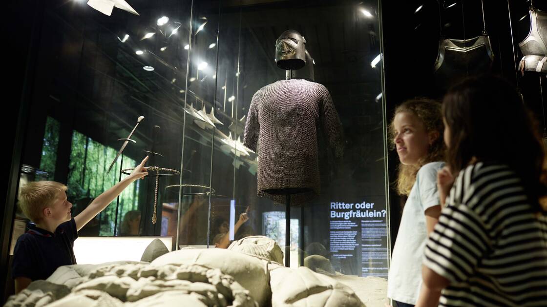 Children stand next to a knight's grave and point to a knight's armour in the showcase