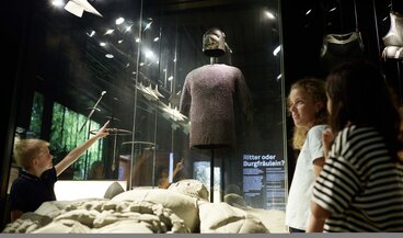 Kinder stehen neben einem Rittergrab und zeigen auf eine Ritterrüstung in der Vitrine