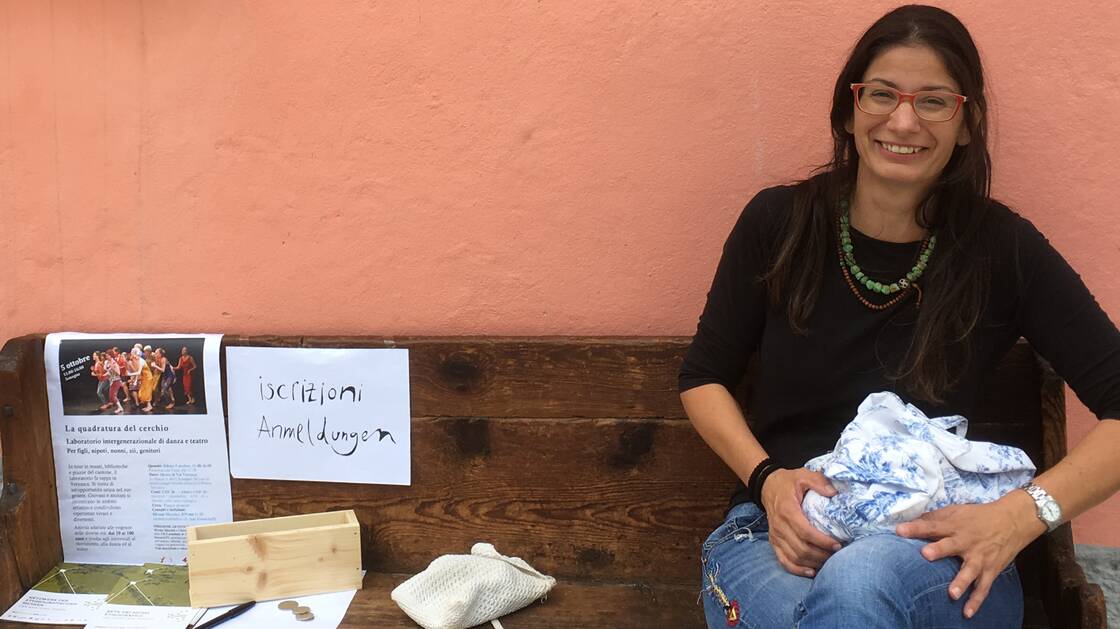 Foto von Veronica Carmine, sie sitzt auf einer Bank vor einer orangenfarbenen Hausmauer