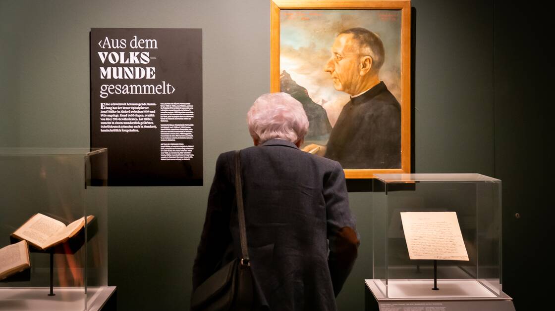An elderly visitor with grey hair stands in front of two display cases and a portrait of hospital priest Josef Müller