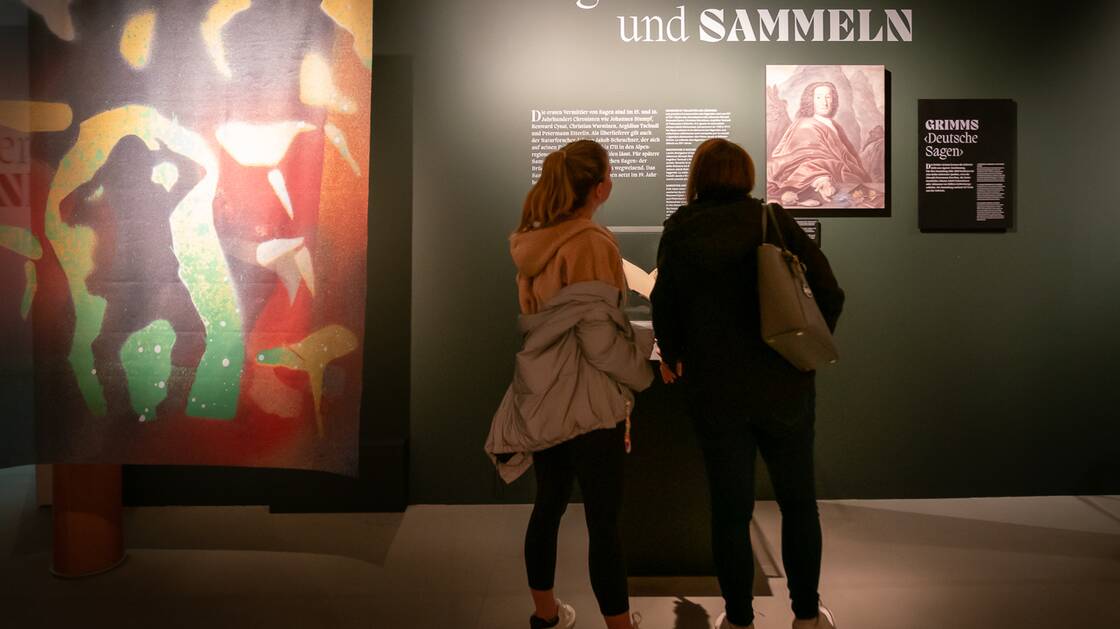 Due visitatori si trovano di fronte a un testo espositivo sul tema "Raccontare e collezionare leggende".